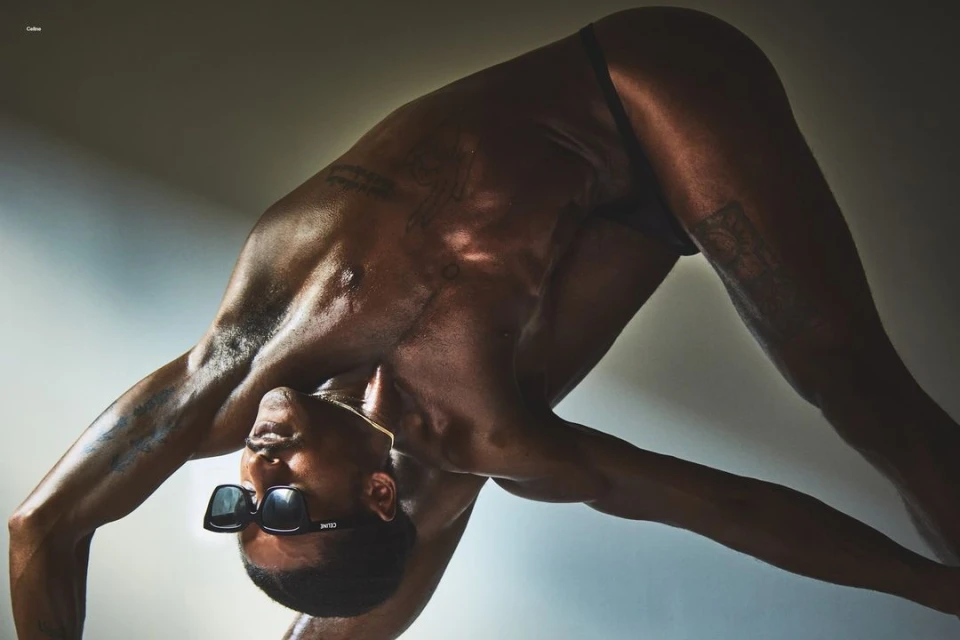 Athletic African American man in sunglasses does a yoga pose indoors with gradient gray and white background lighting the scene.