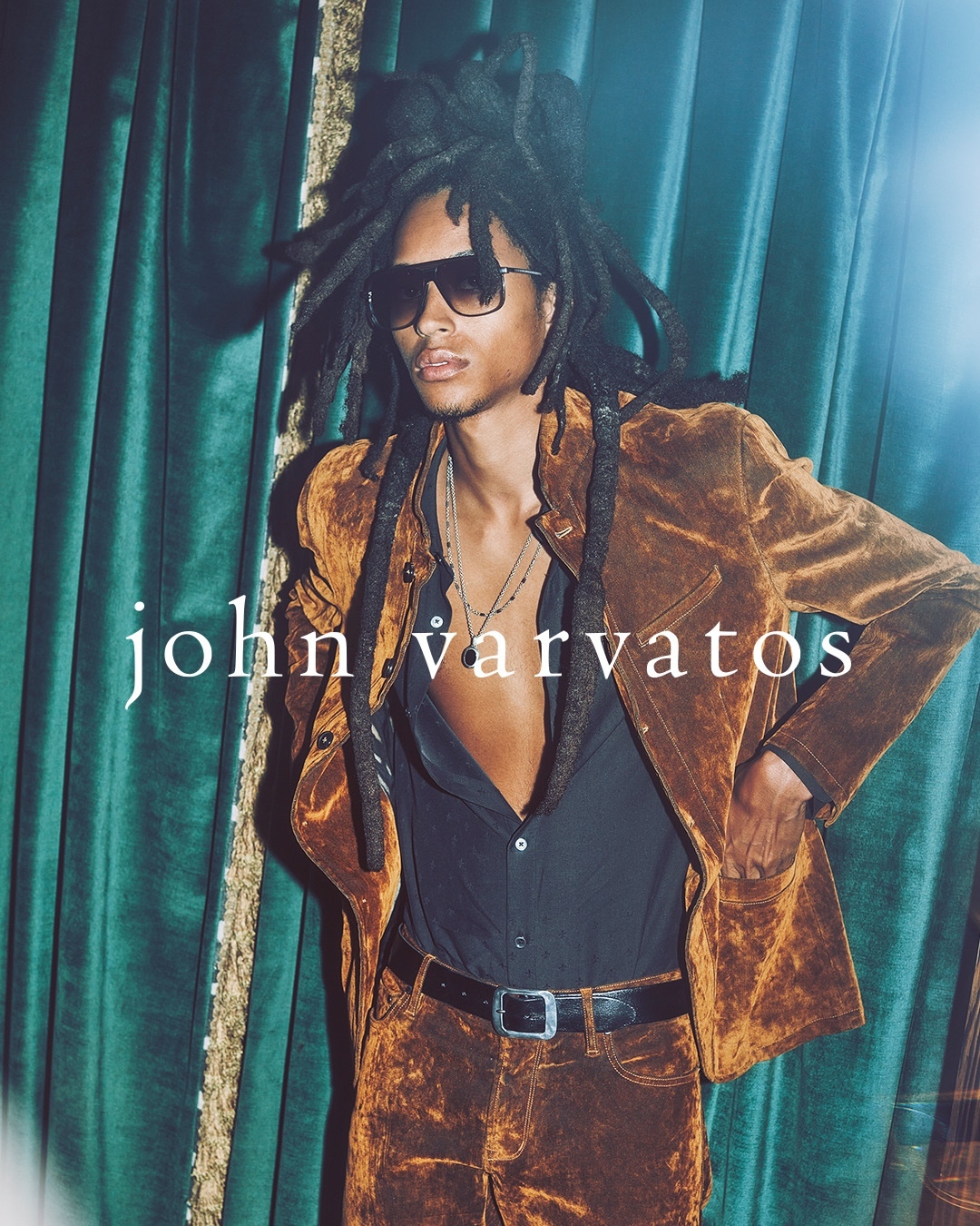 A man in brown velvet suit and sunglasses stands against a green curtain with 'John Varvatos' text overlay. Fashion, style, and portrait.
