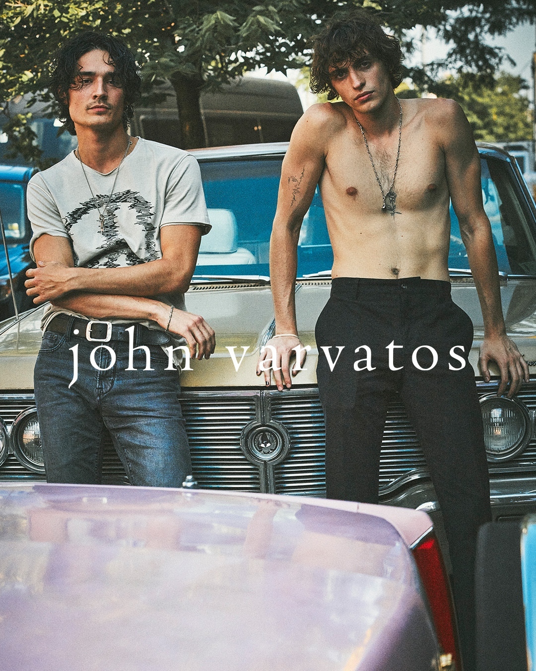 Two male fashion models pose while leaning on classic cars outdoors in the daytime, showcasing a blend of retro and modern style.
