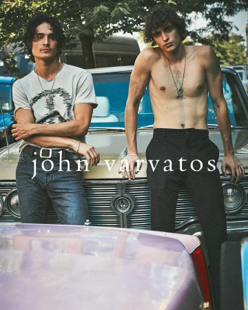 Two male fashion models pose while leaning on classic cars outdoors in the daytime, showcasing a blend of retro and modern style.
