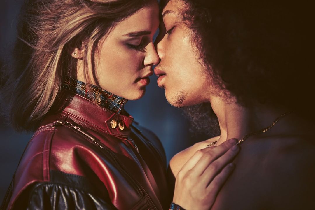 Close-up of a couple, a blonde woman in a leather jacket and a man with curly hair and gold chain, face to face in indoor setting.