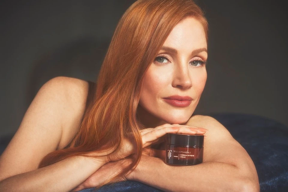 Close-up beauty shot of an adult woman with red hair smiling and resting her chin on her hands with a cream jar in the foreground.
