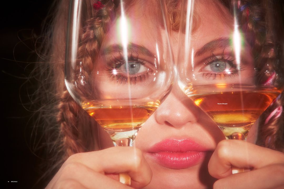 Close-up of an adult female face partially obscured by two wine glasses filled with golden liquid, fair skin, light eyes, and braided hair.