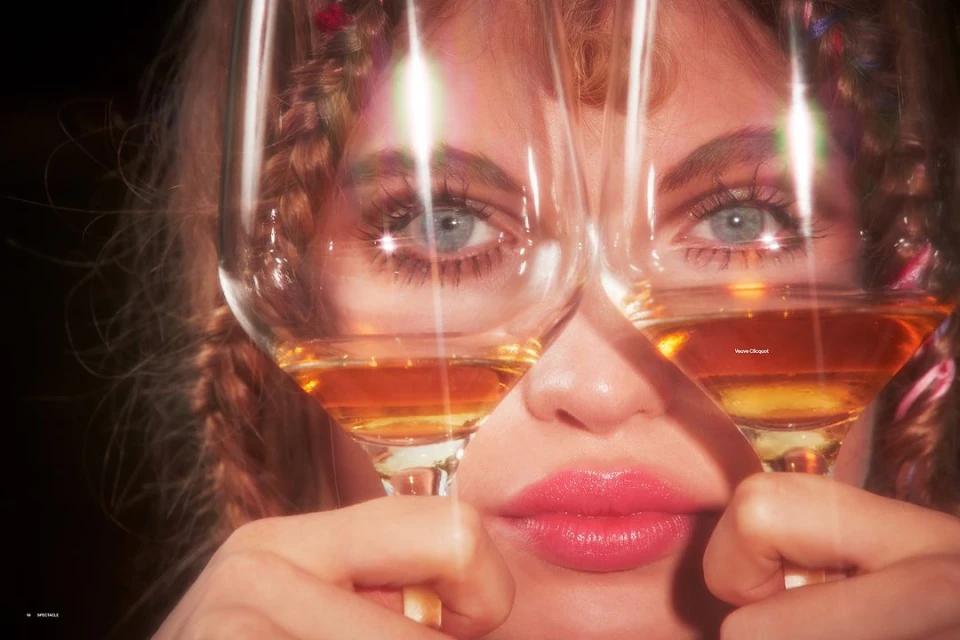 Close-up of an adult female face partially obscured by two wine glasses filled with golden liquid, fair skin, light eyes, and braided hair.