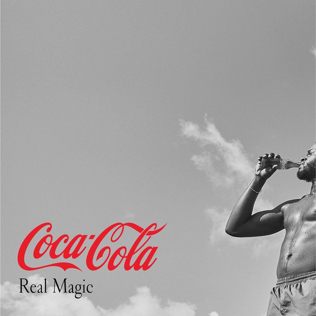 Adult man drinking Coca-Cola against a cloudy sky. The Coca-Cola logo and 'Real Magic' are also visible on the left side.