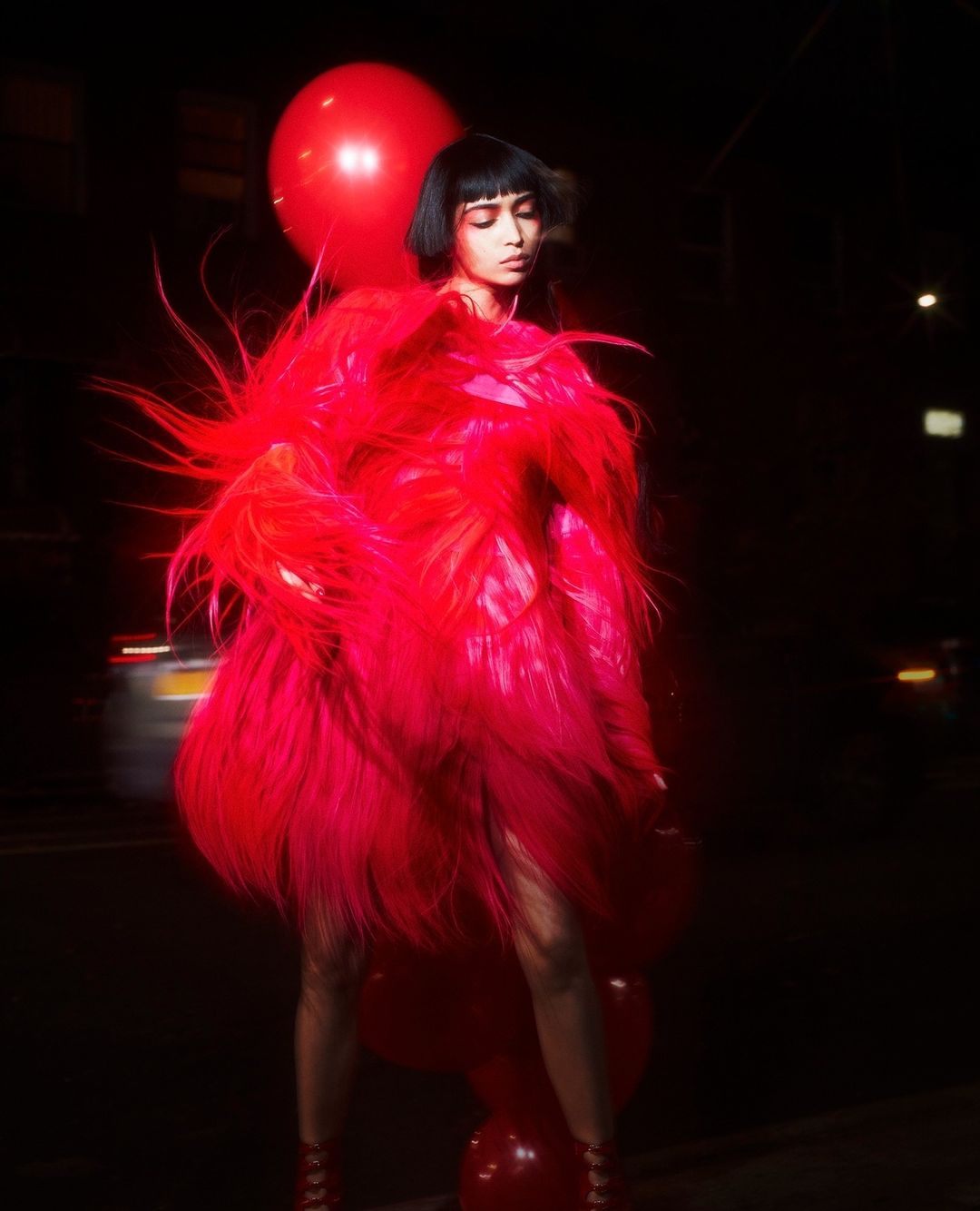 A person in a long red coat stands outside with red balloons against a dark city street backdrop at night.