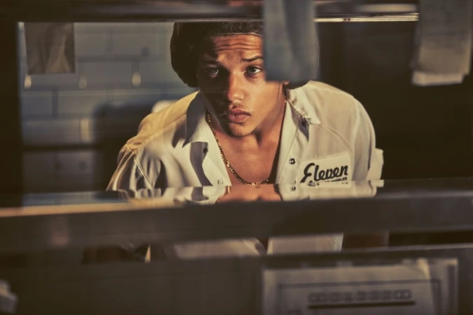 Young male cook wearing white uniform shirt and hairnet looking forward from behind commercial kitchen pass