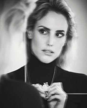 Black and white portrait of a woman reflected in a mirror wearing a dark turtleneck and adjusting a pendant necklace