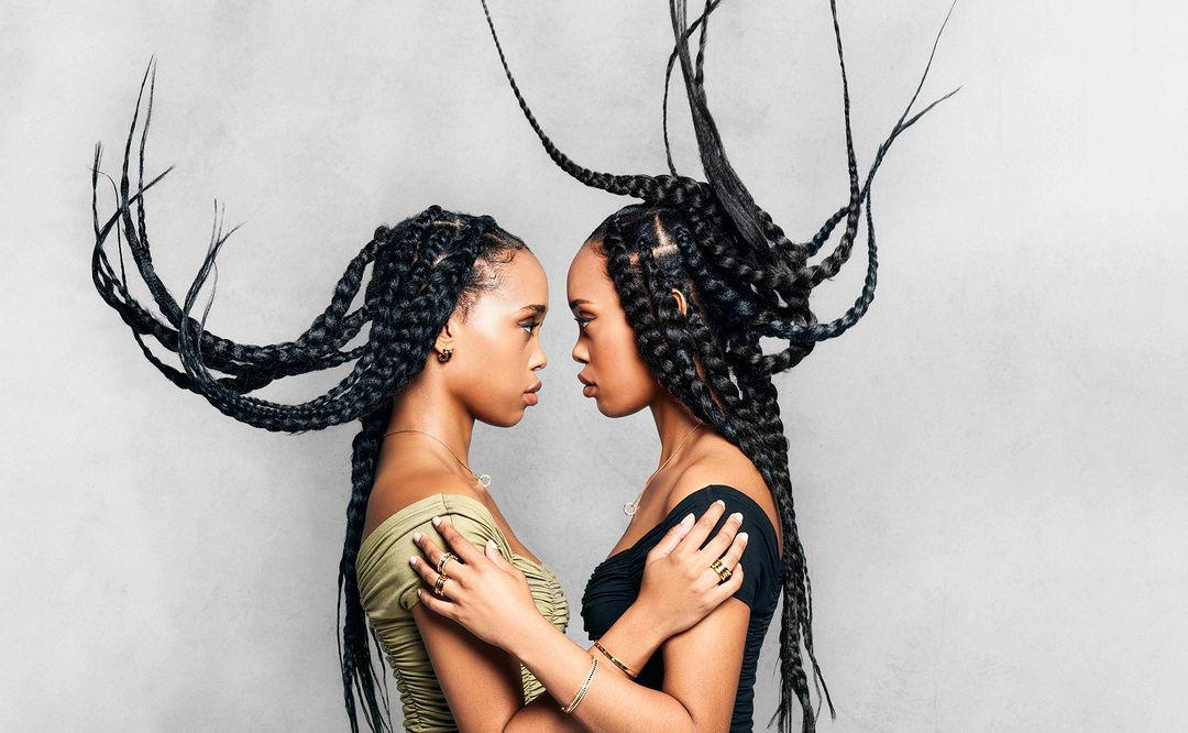 Portrait of two Black women face to face with striking braided hairstyles, accessorized with rings and necklaces, against a grey background.