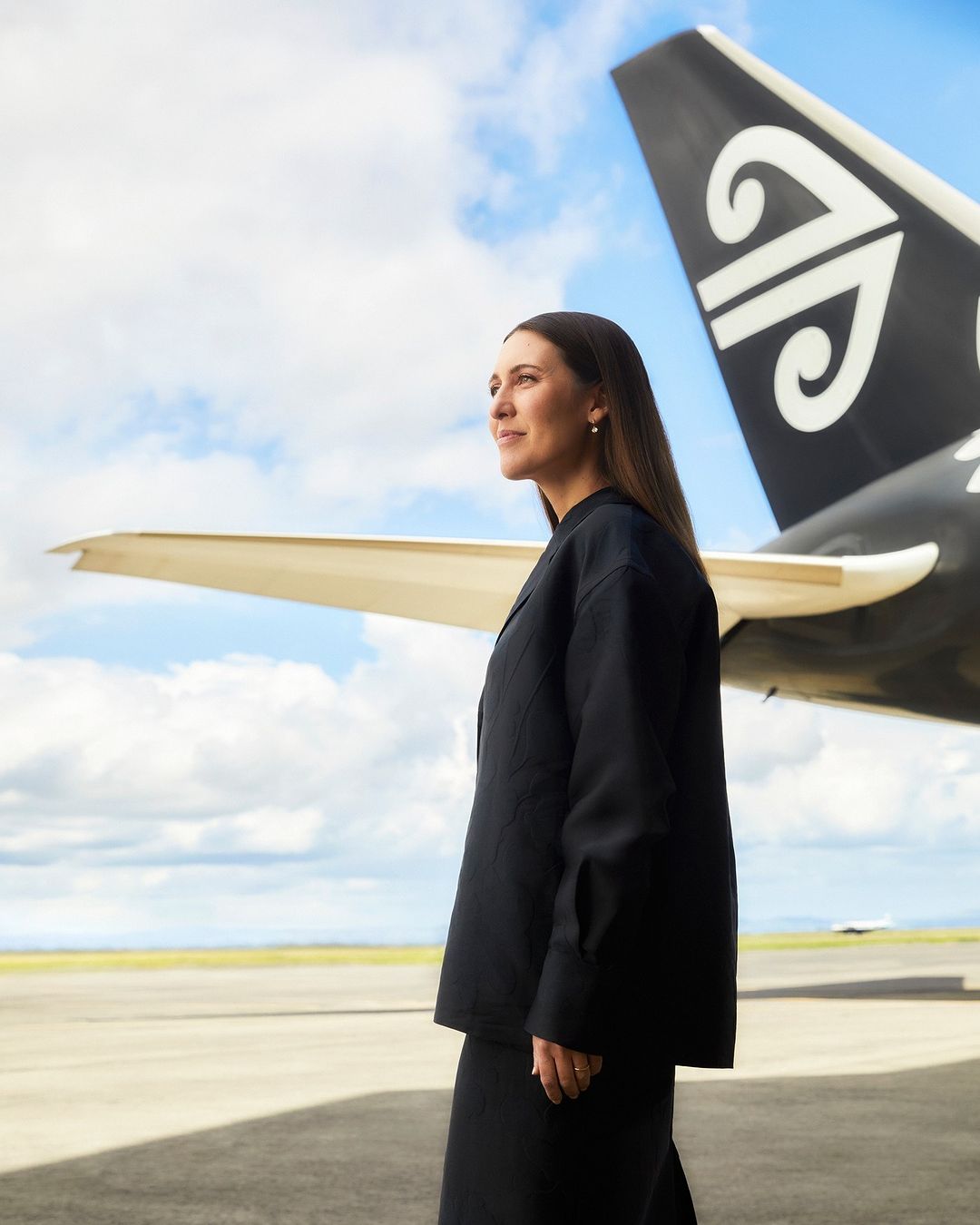 Woman in dark jacket stands by Air New Zealand plane on tarmac under bright cloudy sky, promoting travel and aviation industry.