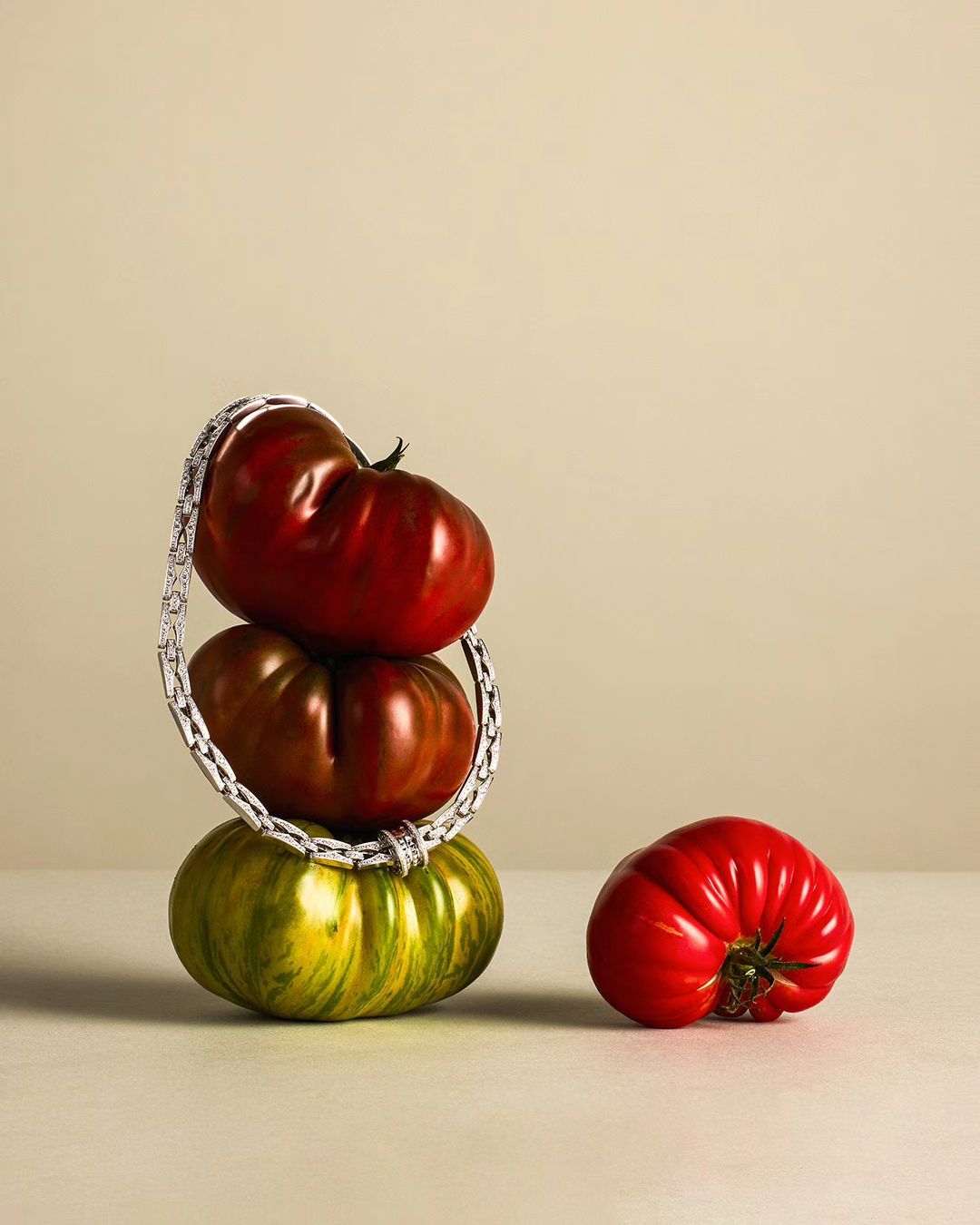 Still life of three heirloom tomatoes stacked with a silver chain jewelry piece draped around them, another tomato on surface.