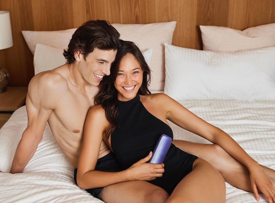 Smiling couple sits on a bed with white linens; the woman holds a small device, showing a cozy, intimate indoor scene.