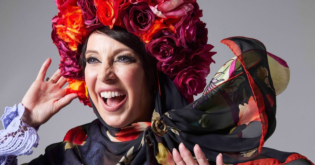 Smiling woman with floral headpiece and colorful scarf posing in front of gray background. Expressive portrait with bold colors.