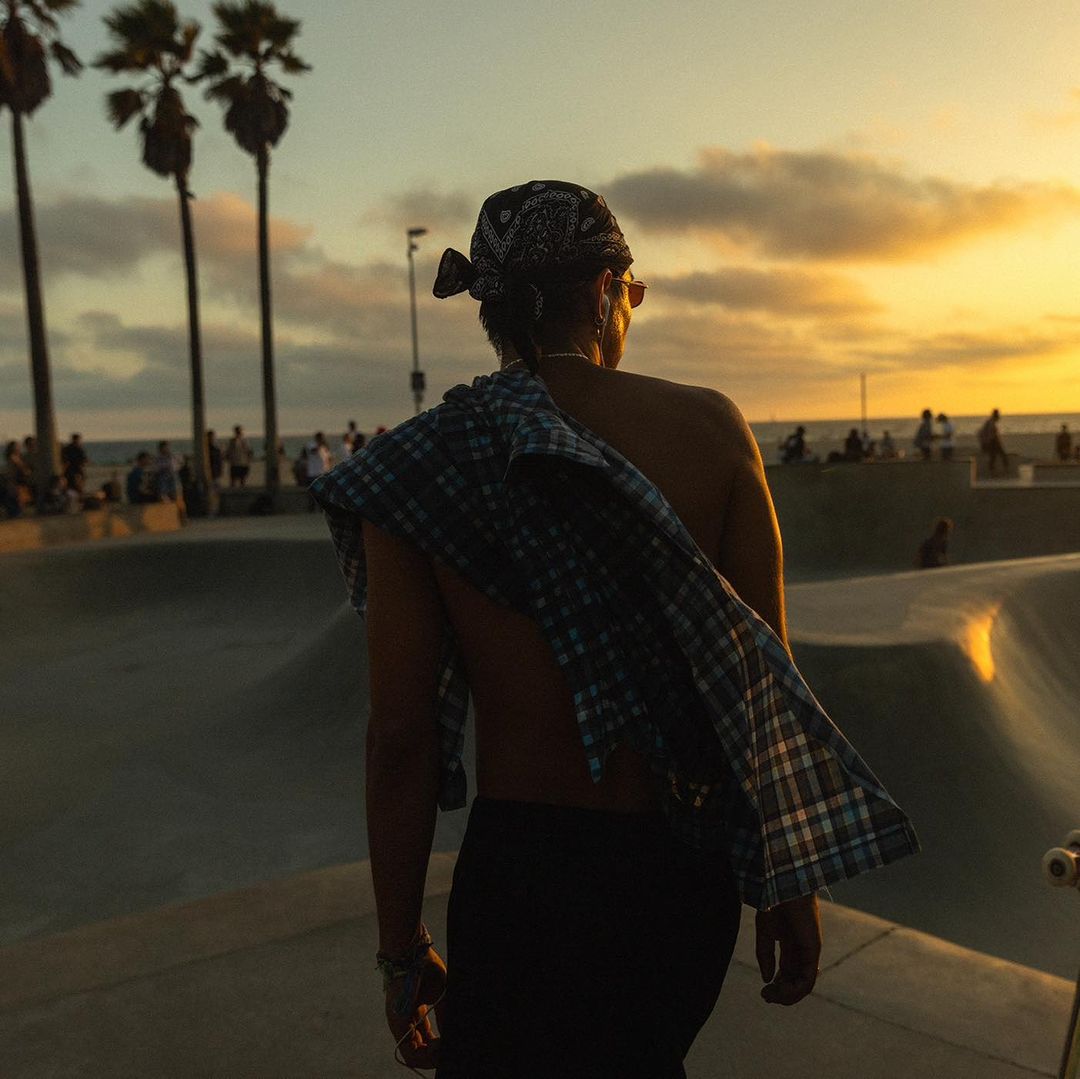 Back view of an adult male in a bandana and black pants at a skatepark during sunset, with palm trees and a crowd visible.