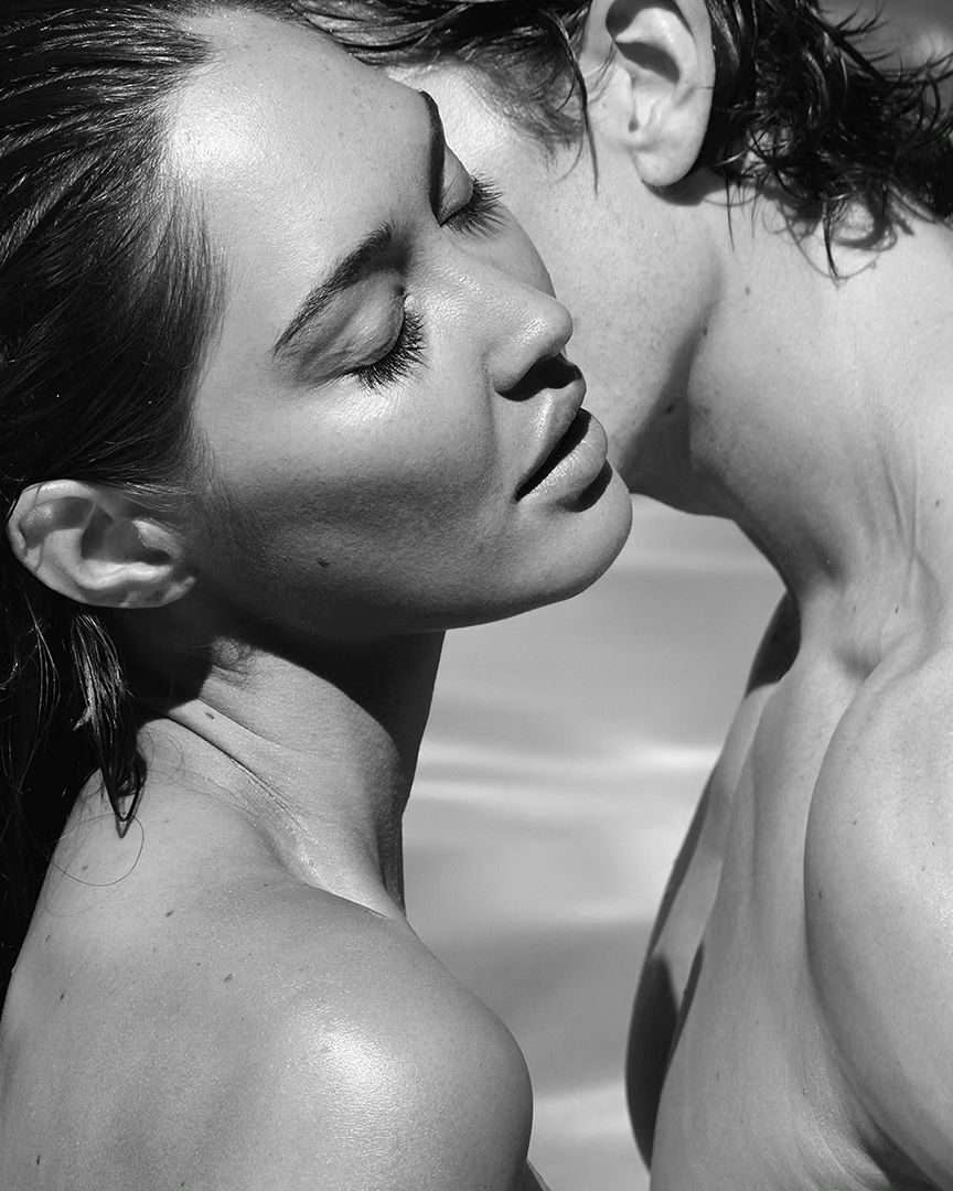 Black and white portrait of an adult woman's face with closed eyes, standing next to an adult man with his face near hers.