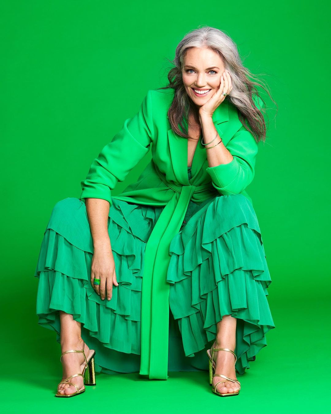 Smiling woman with grey hair in a bright green blazer and ruffle skirt sitting against a green studio backdrop in a fashionable pose.