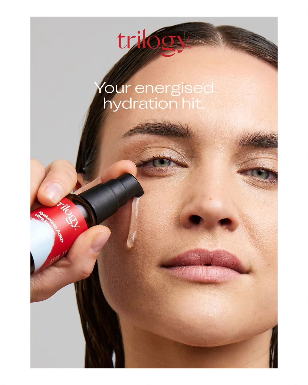Close-up of an adult female applying Trilogy hyaluronic acid serum, with a focus on skincare and beauty, against a plain grey background.