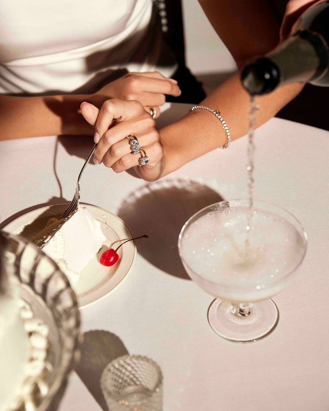 Woman's hands with jewelry reach for glass of sparkling wine as champagne pours from a bottle, alongside a dessert plate with cherry.
