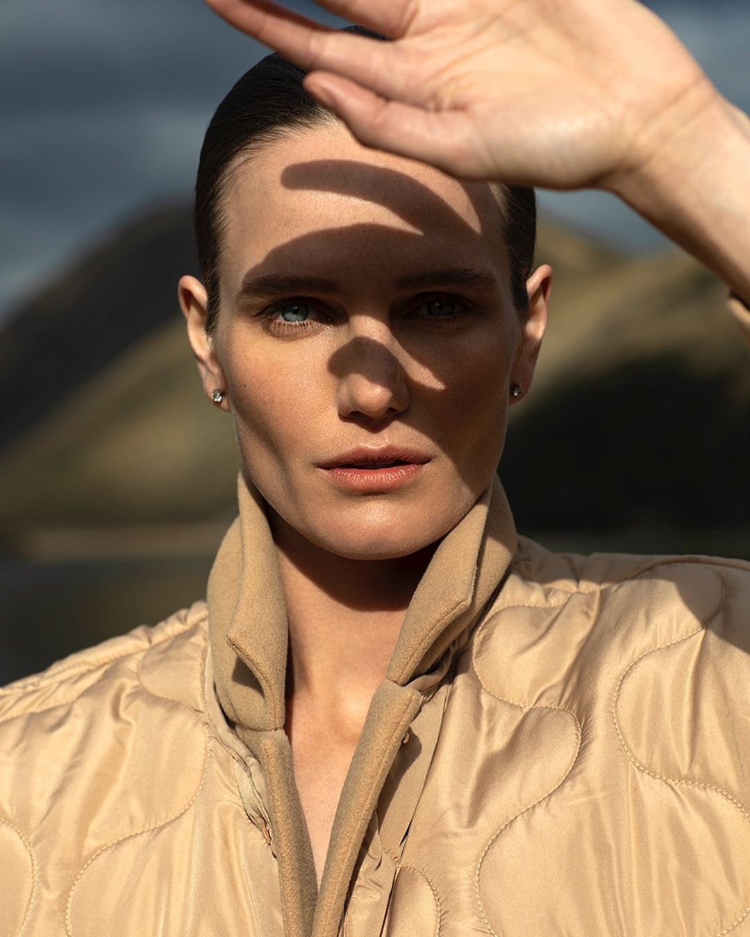 Close-up portrait of a woman shielding her face from sunlight outdoors with mountains and sky in the background.