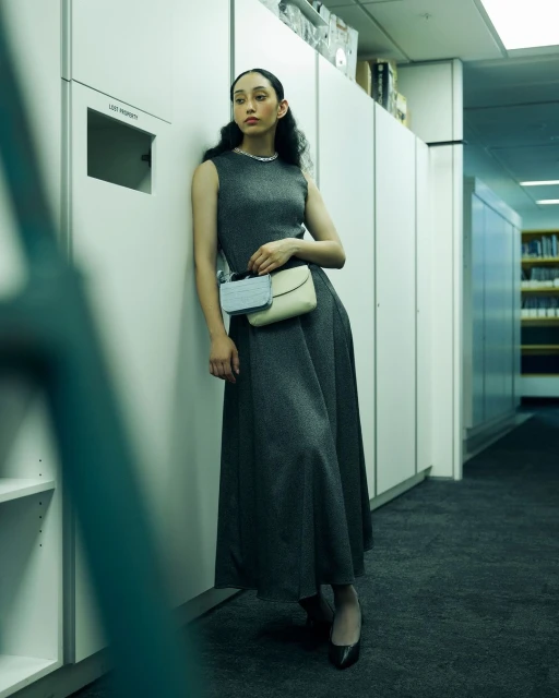 Woman in grey dress and heels poses in hallway with lockers, holding a clutch, fashion shot, indoor lighting.