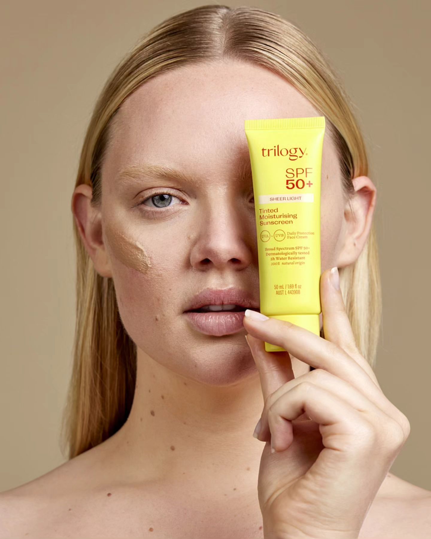 A woman with fair skin holds a yellow tube of Trilogy SPF 50+ sunscreen, face partially covered with sunscreen for skincare.