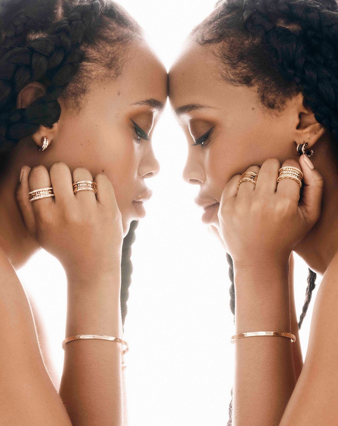 Close-up portrait of two adult women with braided hair, wearing gold jewelry and leaning their heads together against a bright white backdrop.