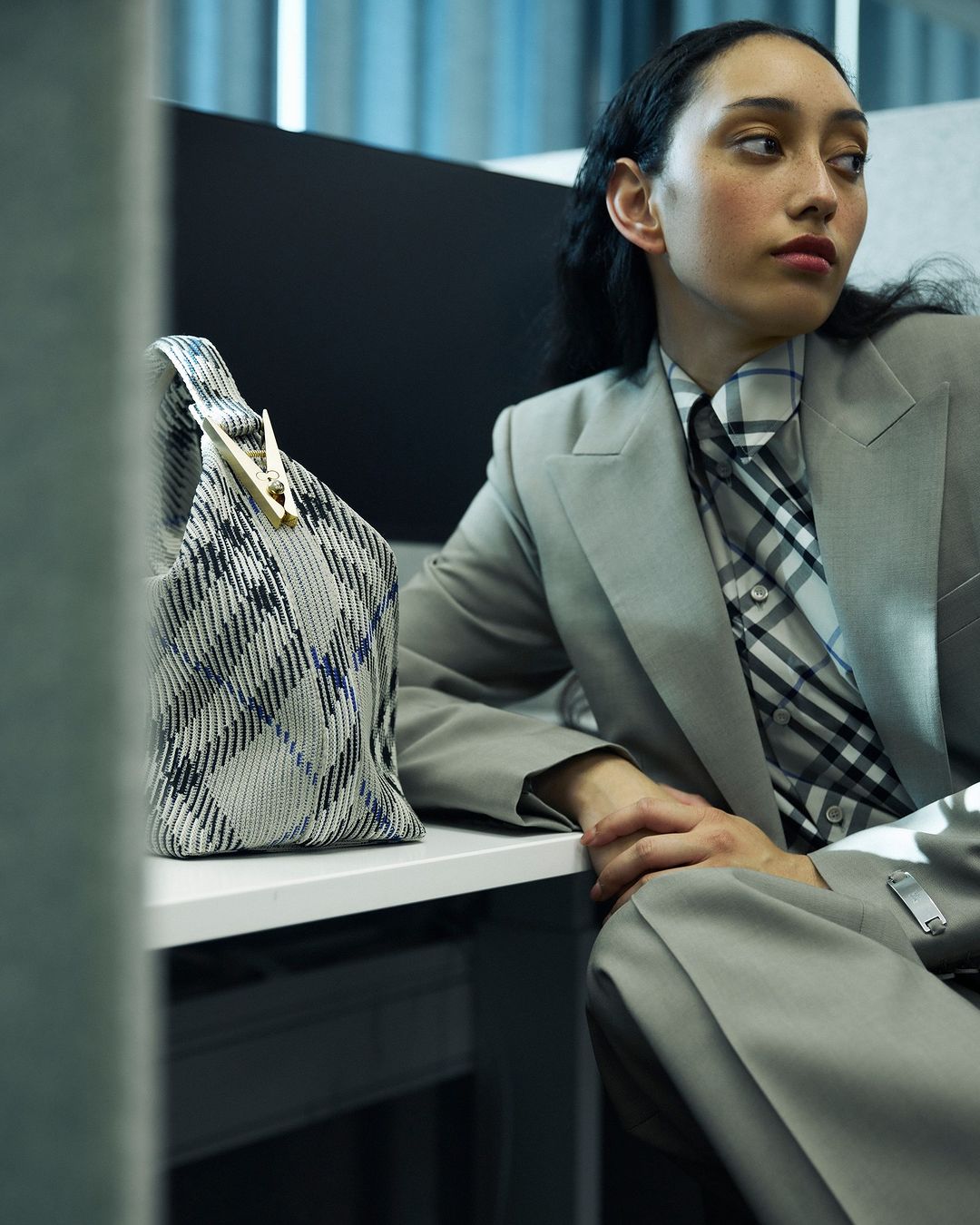 An adult female model sits at a desk wearing a grey pantsuit, with a patterned handbag in the foreground, inside an office space.