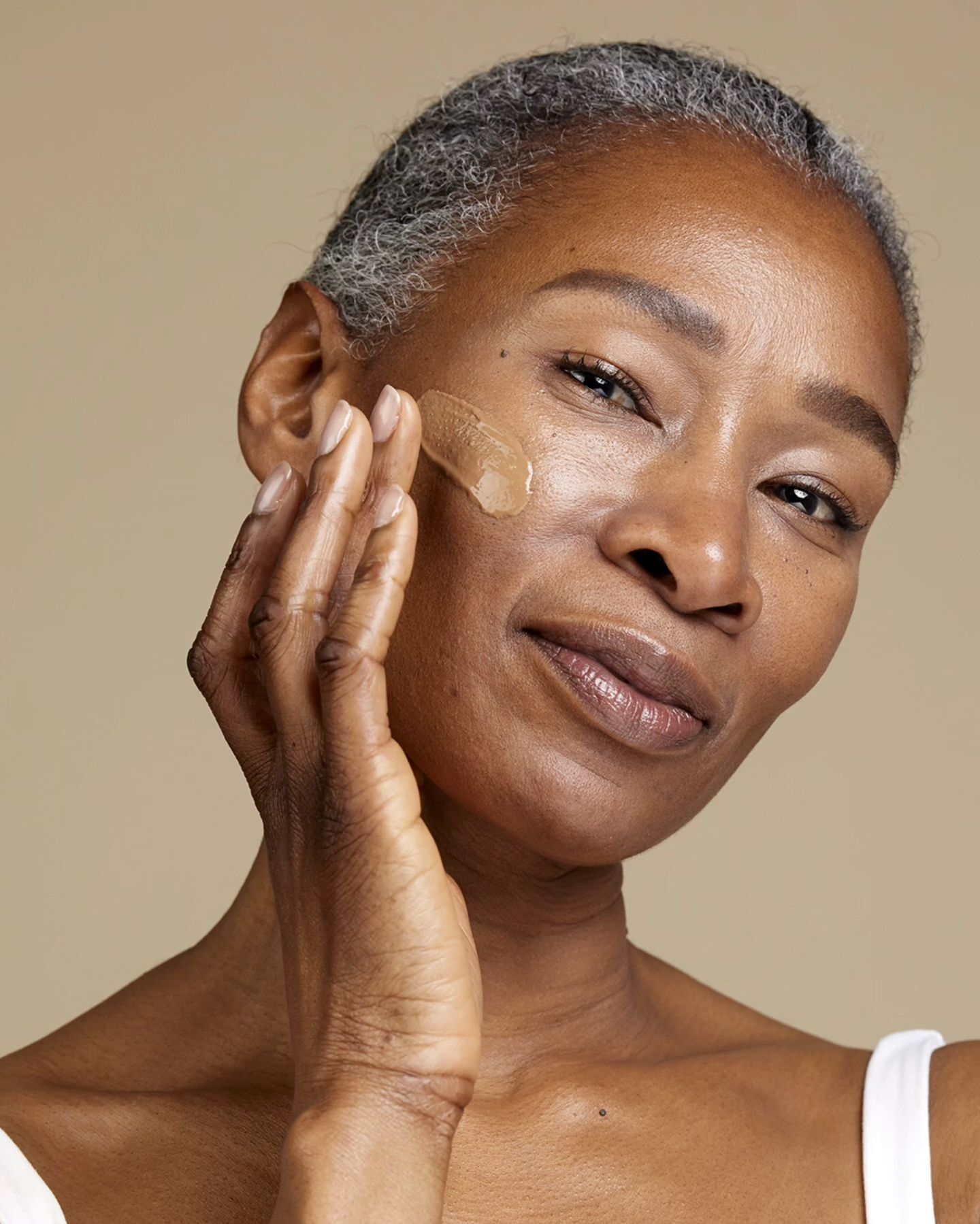Close-up of a mature African American woman applying facial serum to her cheek, showcasing skincare and self-care against a neutral backdrop.