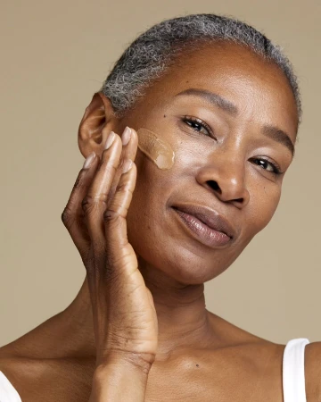 Close-up of a mature African American woman applying facial serum to her cheek, showcasing skincare and self-care against a neutral backdrop.