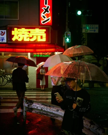People with umbrellas walk through a rainy street in Japan at night, neon signs illuminate the storefronts, creating a vibrant urban scene.