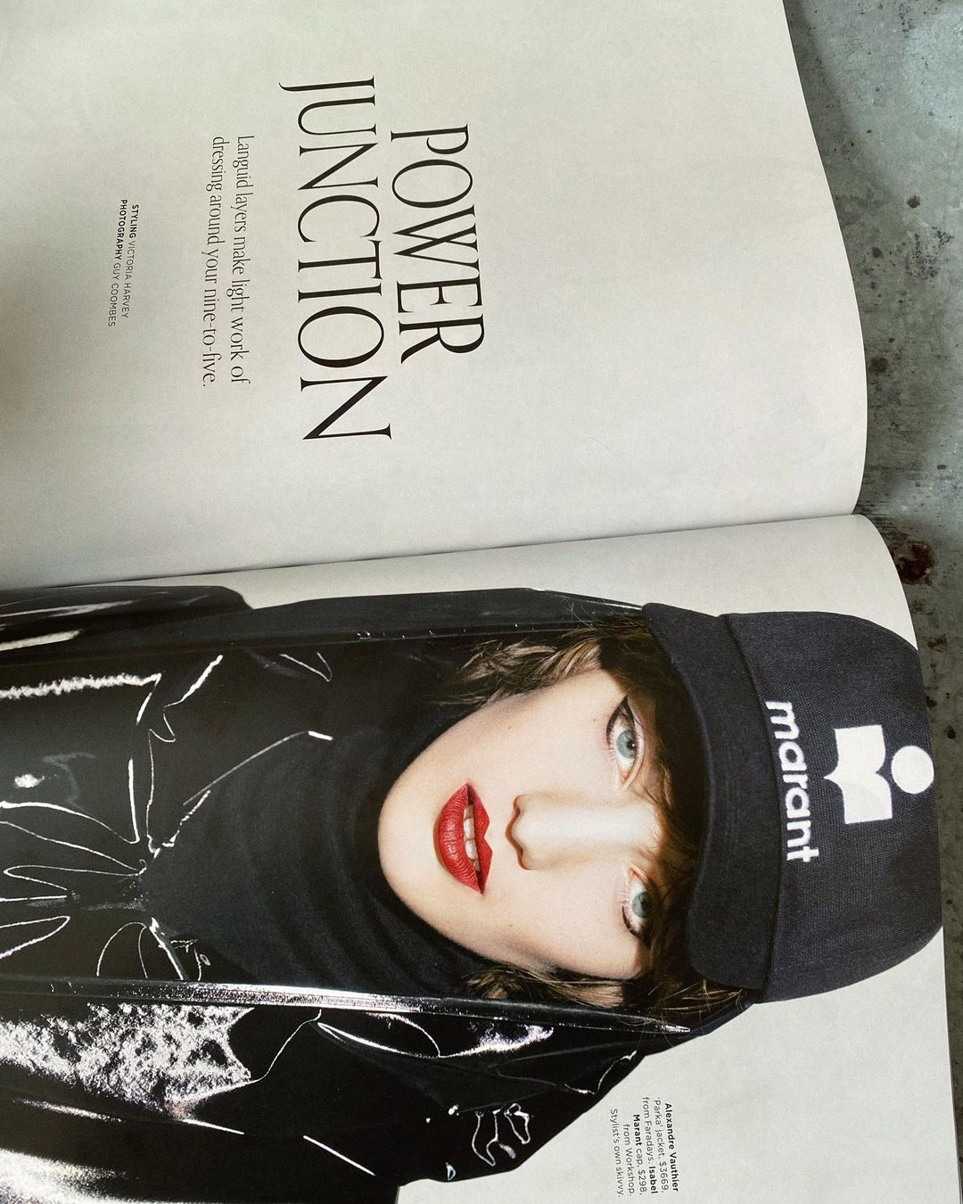 Fashion editorial shot of a young adult female model wearing a black vinyl jacket and a cap with red lipstick on an open magazine.
