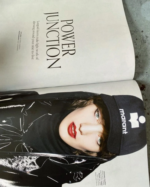 Fashion editorial shot of a young adult female model wearing a black vinyl jacket and a cap with red lipstick on an open magazine.