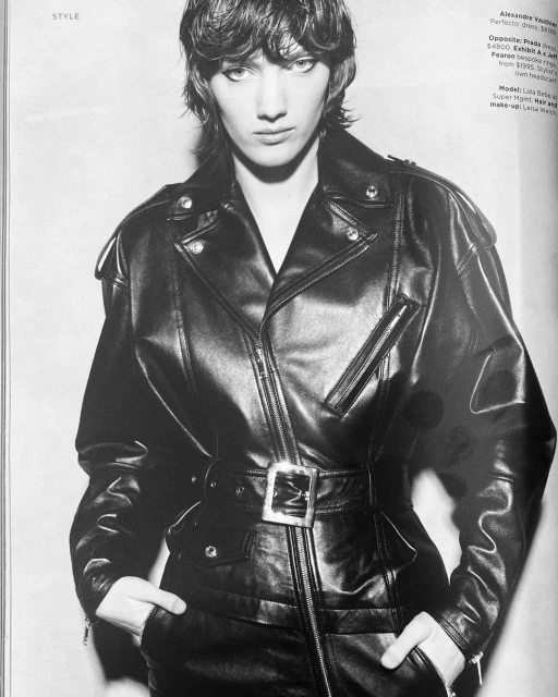 Monochrome fashion shot of a woman with dark hair wearing a leather jacket with a belt, posing indoors against a neutral background.