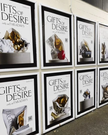 Several framed posters reading 'Gifts of Desire only at Faradays' hang on a wall, each featuring different luxury goods in shopping bags.