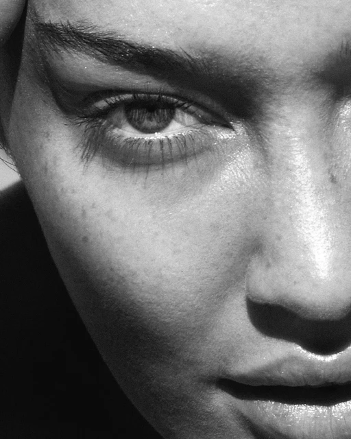 Close-up black and white shot of a woman's face featuring her eye, eyelashes, eyebrow, cheek, nose, and lips, in an indoor setting.