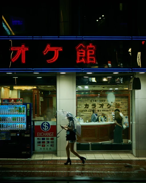 Woman walks under an umbrella in the city at night by a vending machine and karaoke business, with neon lighting reflecting on the wet pavement.