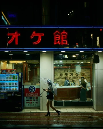 Woman walks under an umbrella in the city at night by a vending machine and karaoke business, with neon lighting reflecting on the wet pavement.