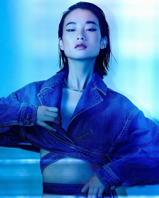An Asian female model with dark hair and striking winged eyeliner wears a denim jacket and a silver choker against a blue backdrop in a studio setting.