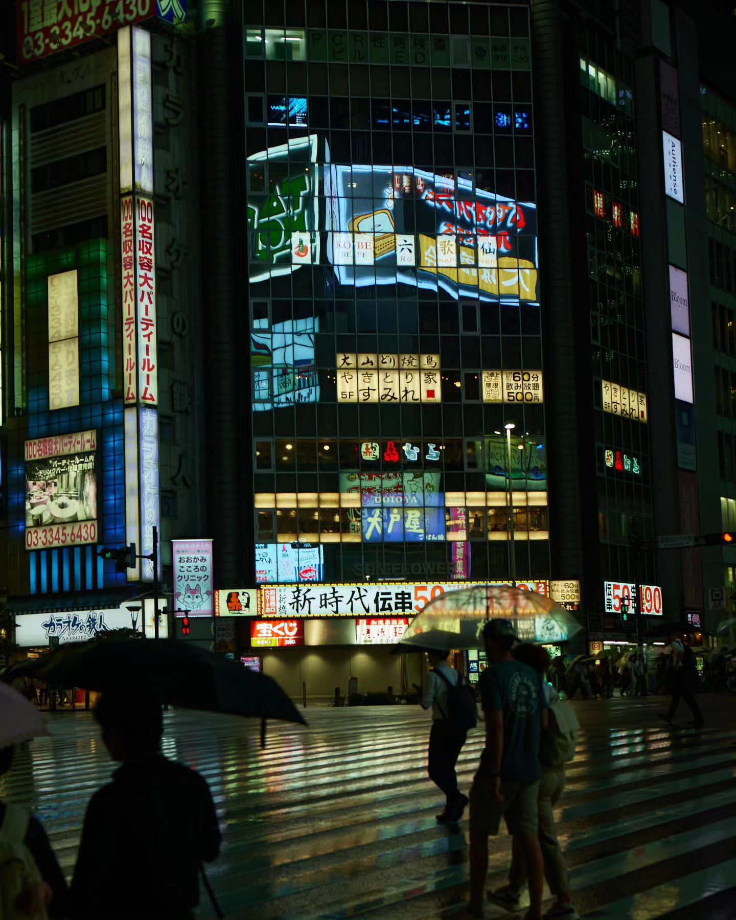Cityscape at night featuring pedestrians with umbrellas crossing a street, surrounded by brightly lit buildings in an urban environment.