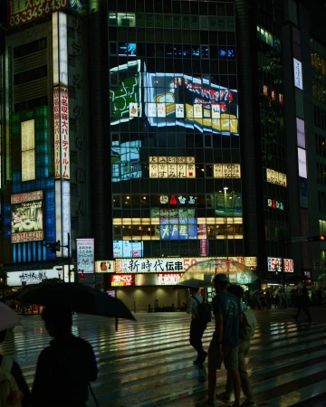 Cityscape at night featuring pedestrians with umbrellas crossing a street, surrounded by brightly lit buildings in an urban environment.