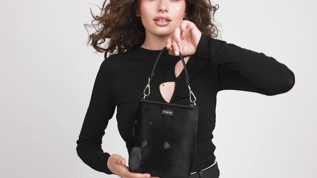 Woman posing in a photo studio holding a black handbag. The model is wearing a long-sleeved black shirt. White background.