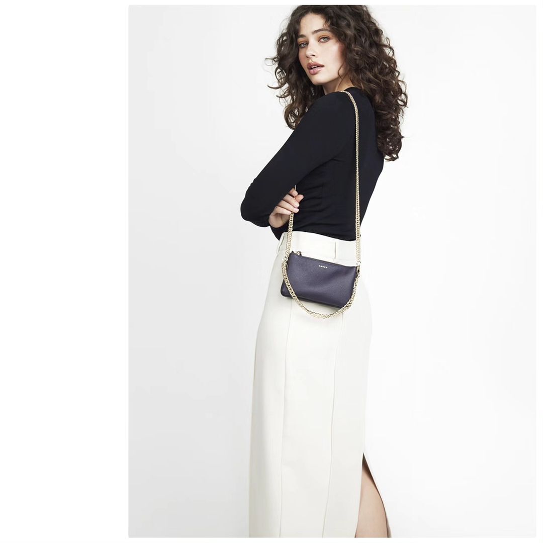 Woman with long curly dark hair wearing a black long-sleeve top and white skirt holding a dark handbag with a chain strap.