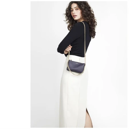 Woman with long curly dark hair wearing a black long-sleeve top and white skirt holding a dark handbag with a chain strap.