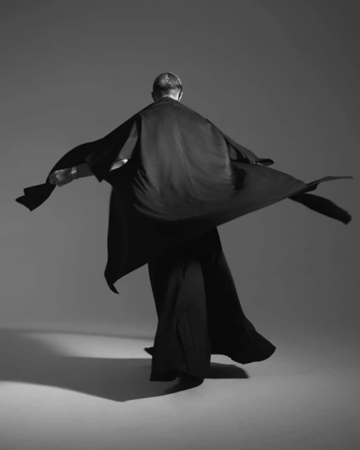 Monochrome portrait from the back of an adult in flowing robe and pants, arms outstretched against a gray studio background.