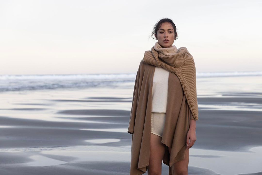 A woman in a beige sweater and cardigan stands on a sandy beach near the ocean under a cloudy sky in a fashion or lifestyle shot.