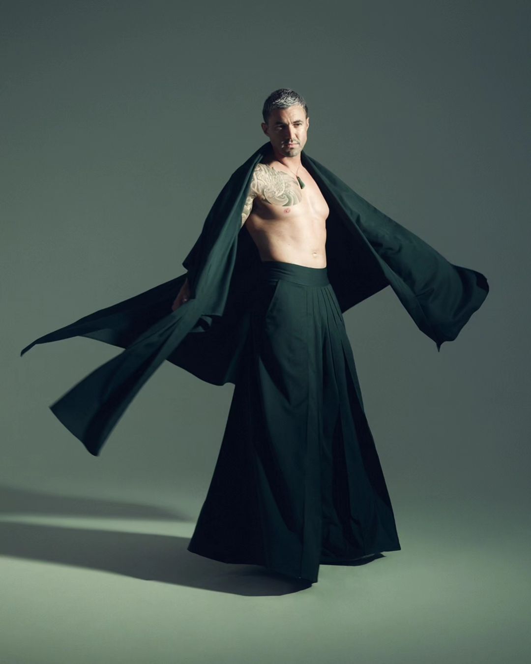 Man with a tattoo on his left arm wearing a long dark robe and dark pants strikes a pose against a neutral grey background in a studio.