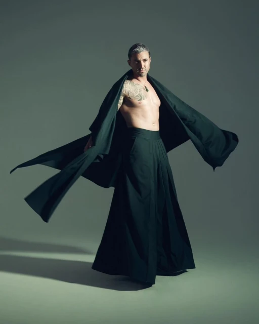 Man with a tattoo on his left arm wearing a long dark robe and dark pants strikes a pose against a neutral grey background in a studio.