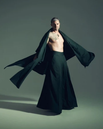 Man with a tattoo on his left arm wearing a long dark robe and dark pants strikes a pose against a neutral grey background in a studio.