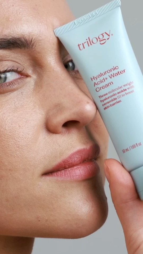 Close-up of a skincare ad with a woman holding a tube of Trilogy Hyaluronic Acid + Water Cream, highlighting beauty and skincare.