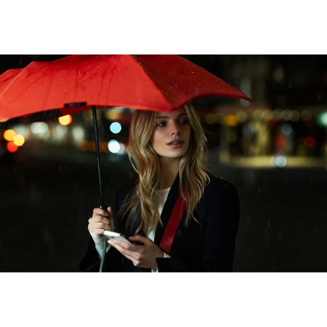 A fair-skinned woman with long blonde hair holds a smartphone under a red umbrella at night on a city street with bokeh lights.